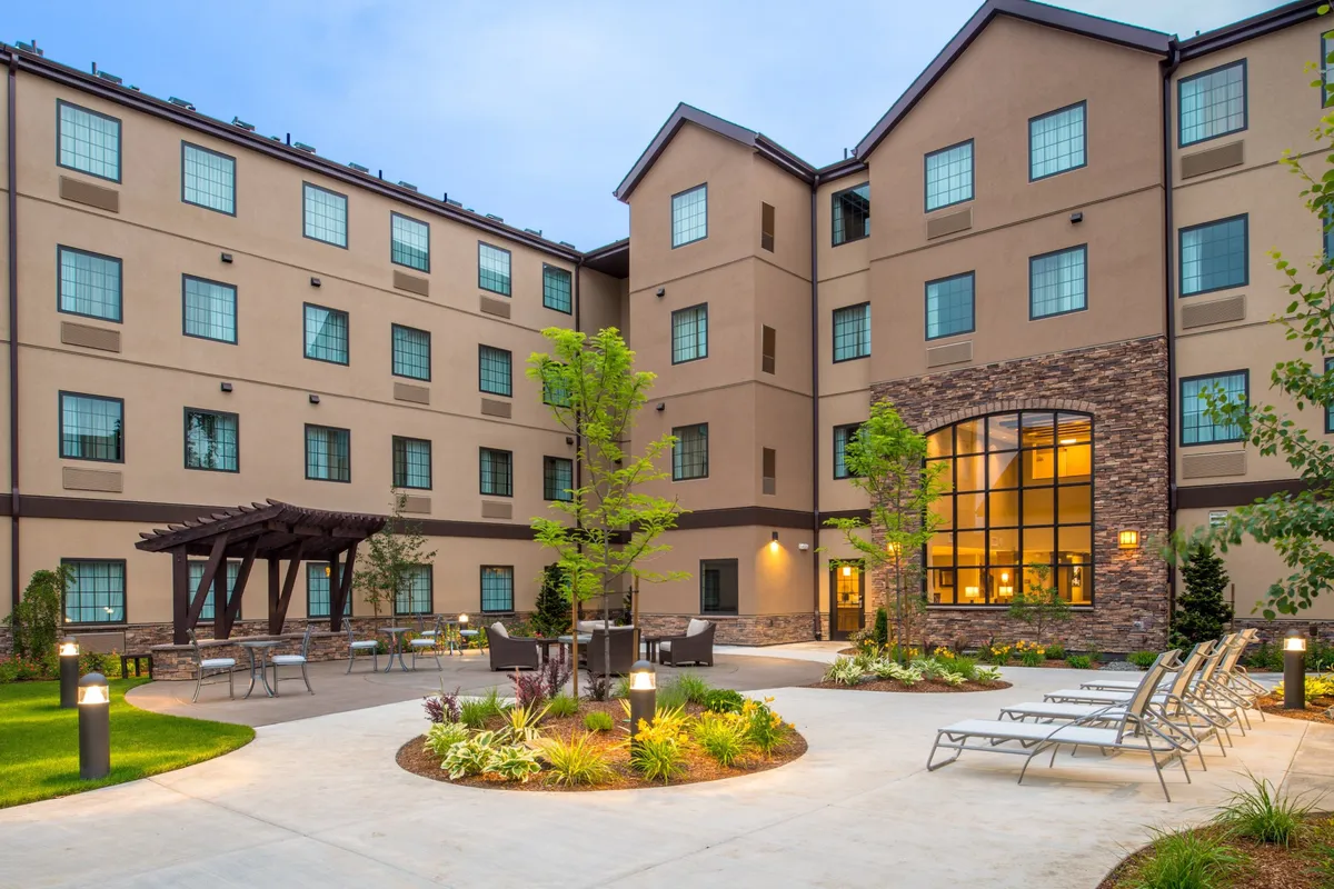 This image displays the hotel's exterior, featuring an inviting outdoor seating area nestled within a landscaped garden. Large windows provide glimpses into the building's interior, while the sunset casts a warm, welcoming glow. The space is ideal for relaxing or gathering outdoors, with greenery and open skies hinting at nearby adventures.