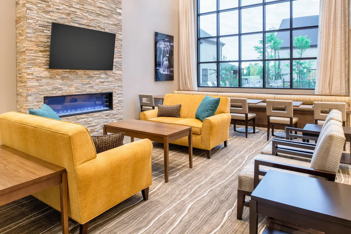 A welcoming living area furnished with yellow sofas, wooden tables, a large window letting in daylight, and a stone feature wall. The setup invites guests to relax, watch TV, socialize, or enjoy a quiet moment, capturing the suite's inviting and flexible living space.