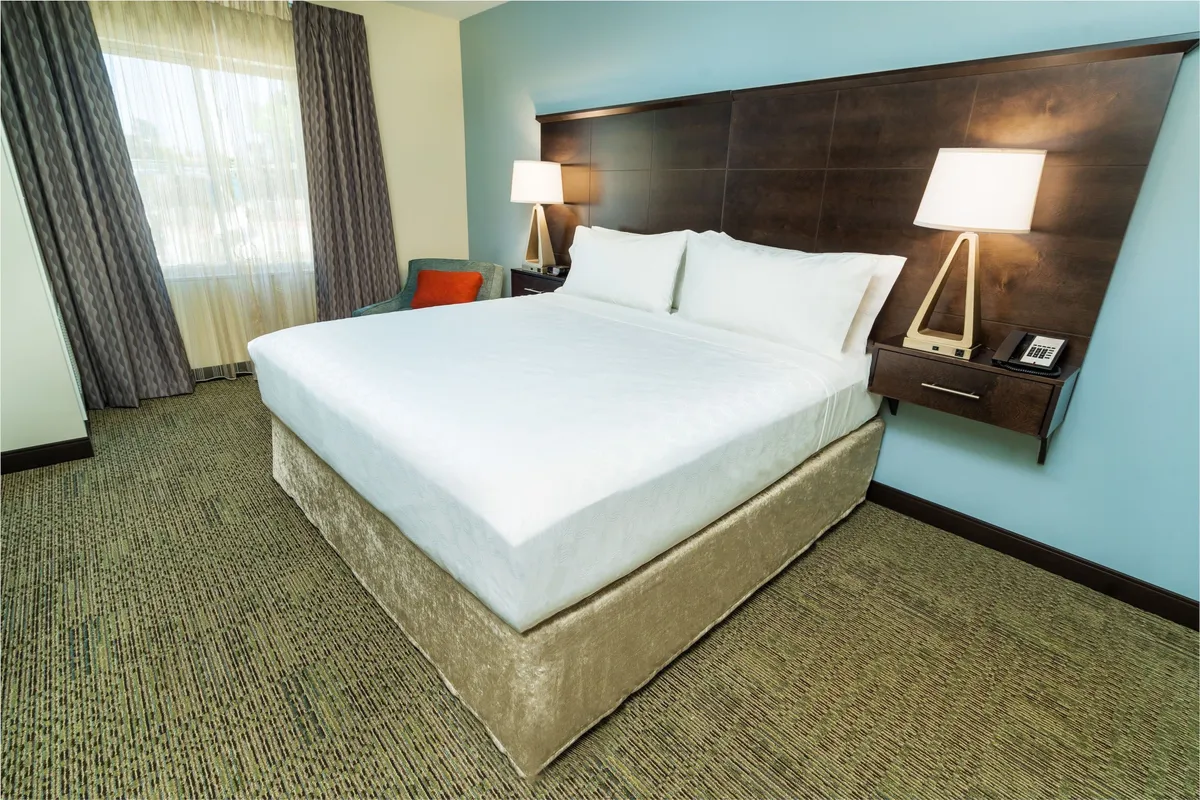 A calm and serene king bedroom featuring a comfortable bed with soft, layered bedding. The space includes a nightstand, window with gentle natural light, and tasteful curtains, emphasizing an atmosphere designed for restful sleep and relaxation.
