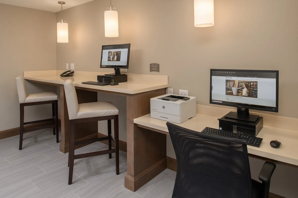 A modern business center with two desktop computers, a printer, and bar-style seating. The space is quiet, well-lit, and arranged for both individual work and small group collaboration. Clean lines and a contemporary aesthetic promote focus and productivity.
