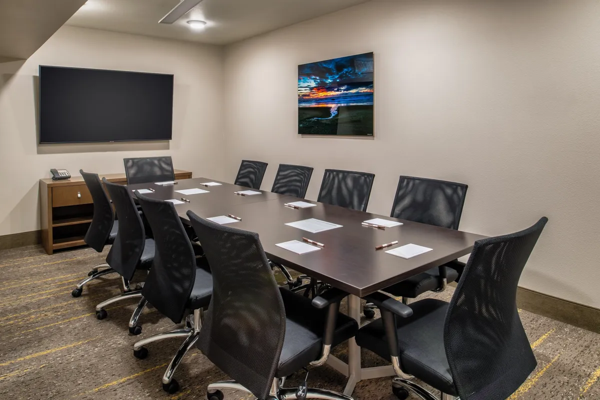 A spacious, well-lit conference room featuring a large table surrounded by comfortable chairs, a prominent TV screen for presentations, and tasteful artwork on the walls. The setup creates a professional environment, ideal for business meetings, brainstorming sessions, or collaborative work.