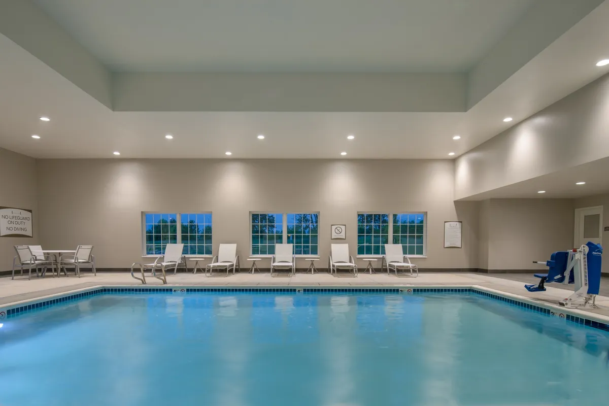 A bright, inviting indoor swimming pool area with clear blue water, several lounge chairs arranged poolside, and large windows letting in natural light. The scene suggests relaxation and family fun, with ample space for both swimming and poolside gatherings.